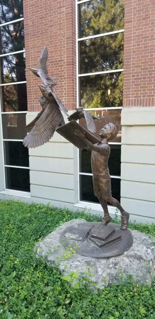 A metal sculpture of a child holding a book with birds flying out of it.