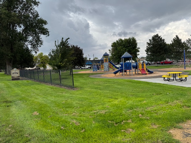 A picture of a city park in the summer with large playstructures and picnic tables in the background